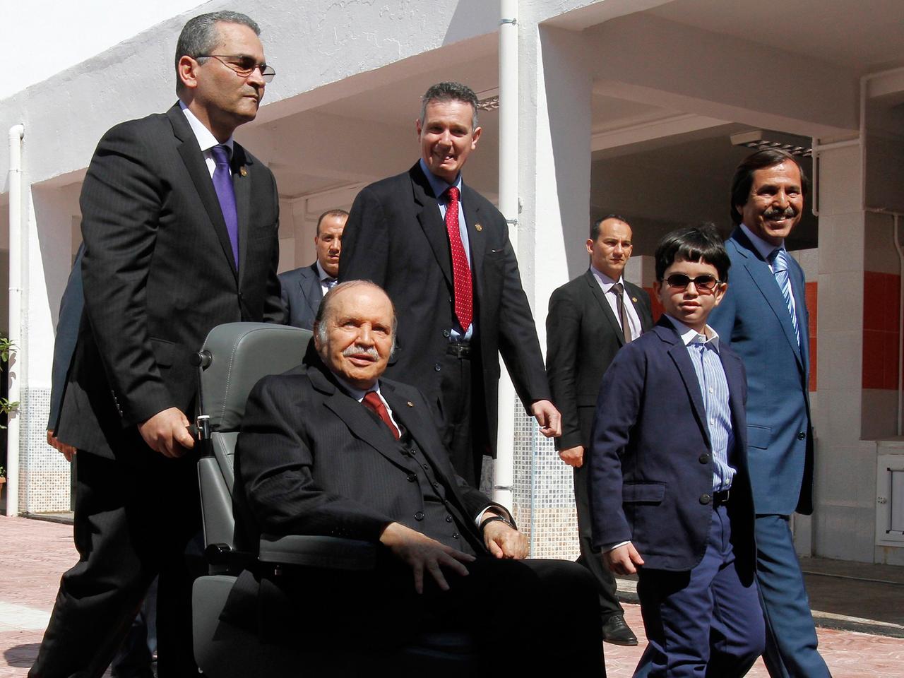 Abdelaziz Bouteflika wird im Rollstuhl von einem Mann geschoben, hinter ihm weitere Männer und ein Junge