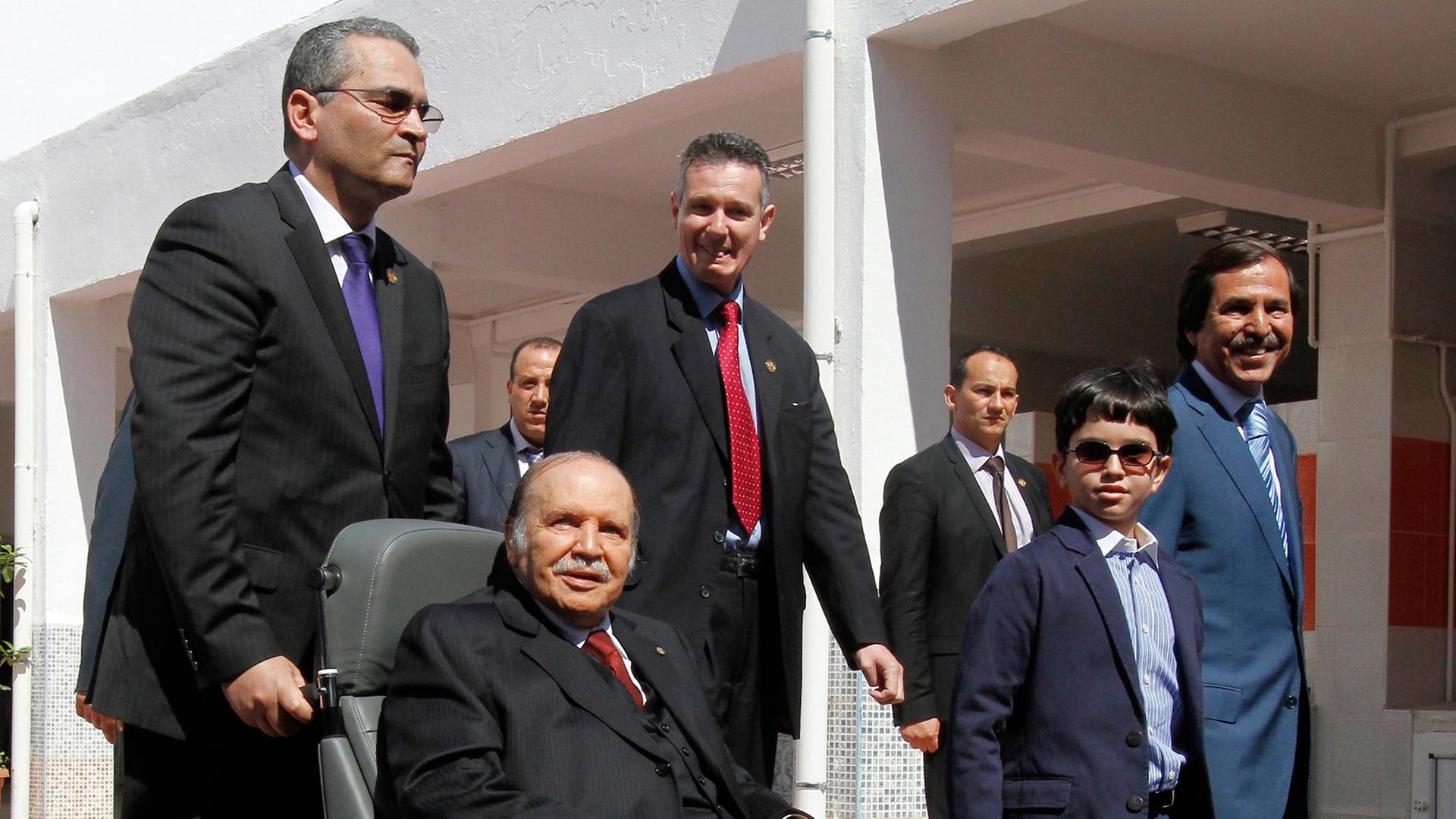 Abdelaziz Bouteflika wird im Rollstuhl von einem Mann geschoben, hinter ihm weitere Männer und ein Junge
