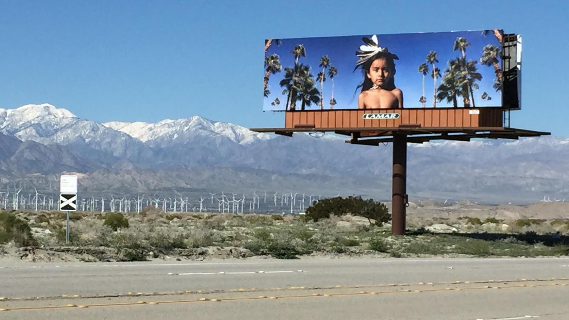Auf dem Bild ist die Arbeit "Winka & The Windmills" der Künstlerin Cara Romero zu sehen. Auf einem Billboard ist das Foto eines kleinen Jungen zu sehen. Er trägt Federkopfschmuck, ein Tuch um die Lenden und eine Sonnenbrille - hinter ihm ragen weiße Windmühlen in den dunkelblauen Himmel.