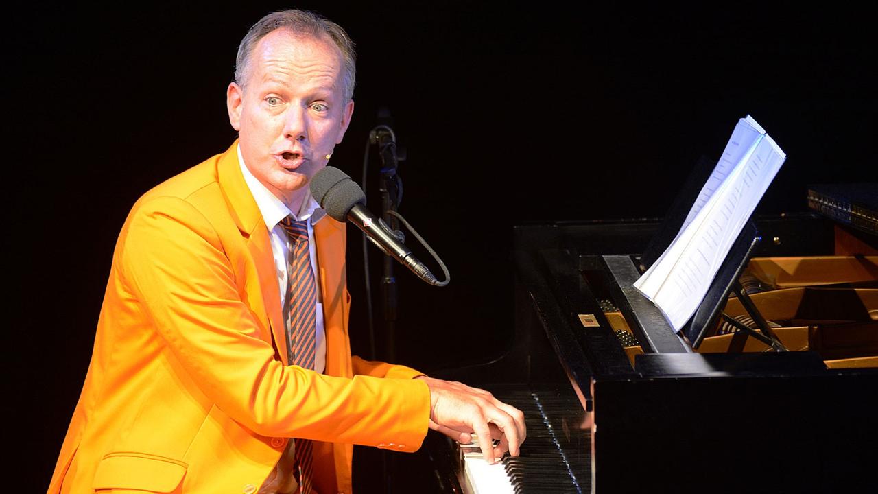 Johannes Kirchberg in orangenem Anzug sitzt am Klavier, spielt und singt