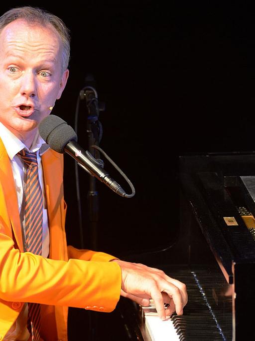 Johannes Kirchberg in orangenem Anzug sitzt am Klavier, spielt und singt