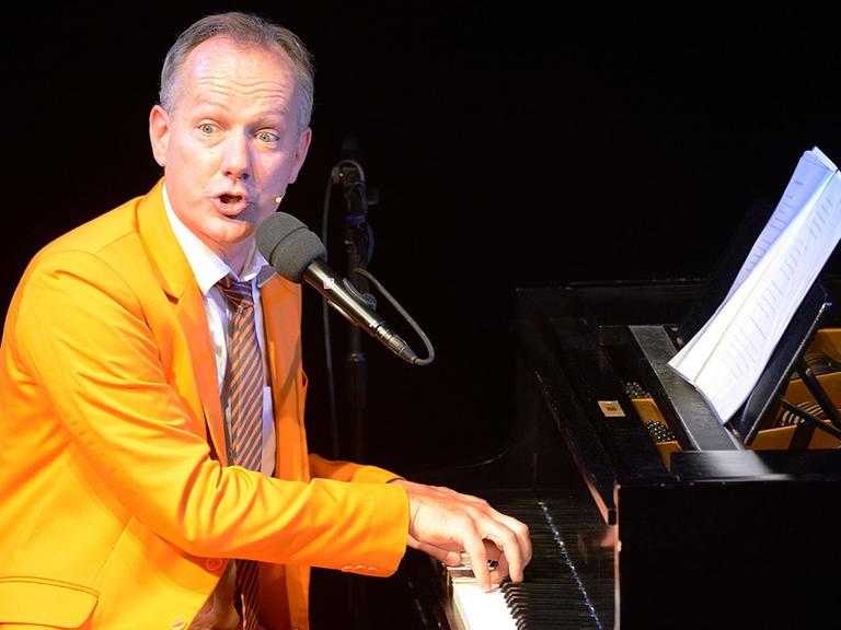 Johannes Kirchberg in orangenem Anzug sitzt am Klavier, spielt und singt