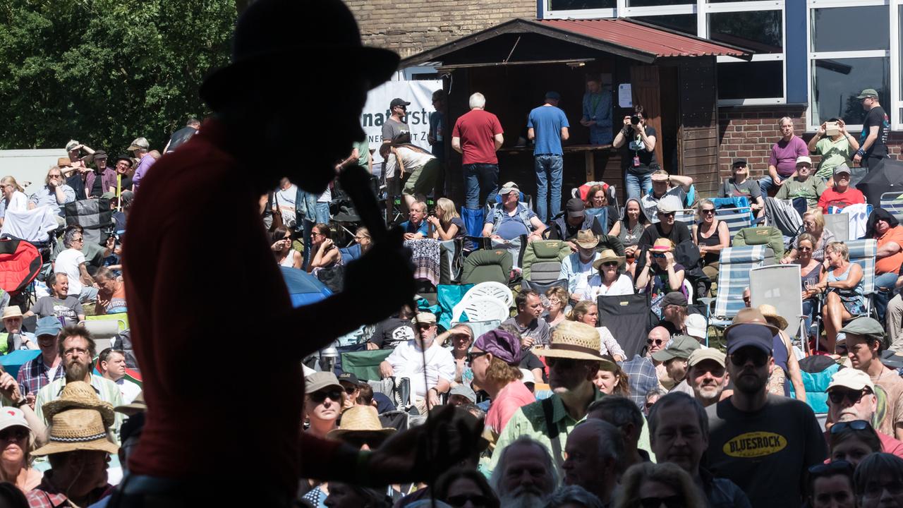 Ein Mann im Profil singt in ein Mikrofon. Im Hintergrund sitzt das Publikum auf Stühlen in der Sonne.