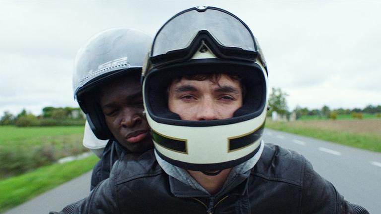 Die beiden Schauspieler Fionn Whitehead und Stéphane Bak sitzen mit Helmen auf einem Motorrad.
