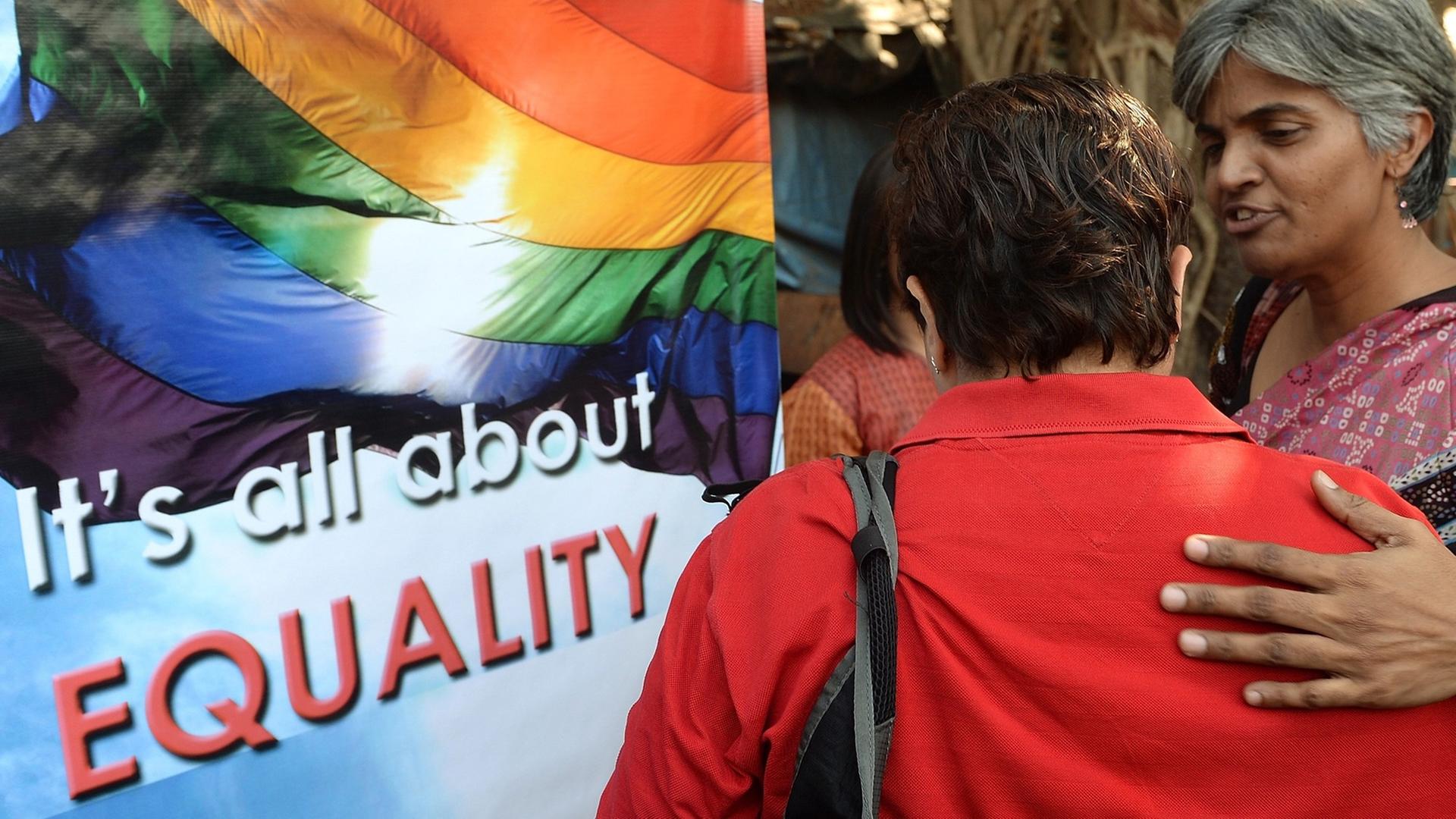 Vor einem Plakat mit einer Regenbogenflagge, dem Symbol für die Schwulen- und Lesbenbewegung, mit der Aufschrift "It's all about Equality" stehen zwei Frauen, eine davon mit dem Rücken zur Kamera, und betrachten es. Aufgenommen wurde das Bild bei einer Demonstranten für die Rechte Homosexueller in Mumbai am 11. Dezember 2013.