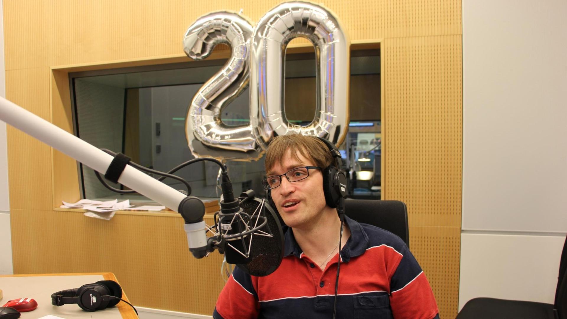 Ein Mann spricht im Radiostudio in ein Mikrofon, während im Hintergrund zwei silberne Ballons in Form einer 20 zu sehen sind