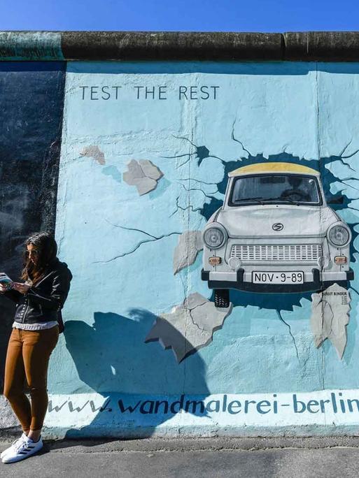 Eine junge Frau steht lesend vor der Berliner Mauer. Darauf ist eine Wandmalerei: ein Trabi, der die Mauer durchbricht.