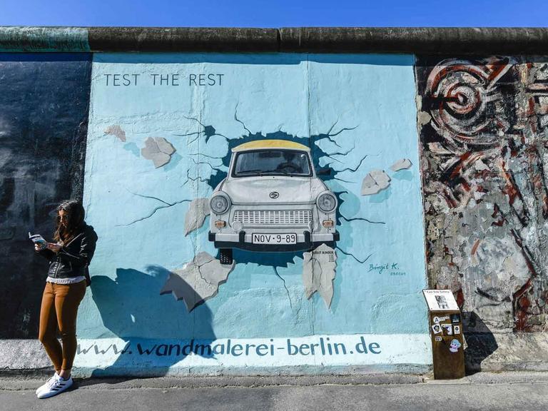 Eine junge Frau steht lesend vor der Berliner Mauer. Darauf ist eine Wandmalerei: ein Trabi, der die Mauer durchbricht.