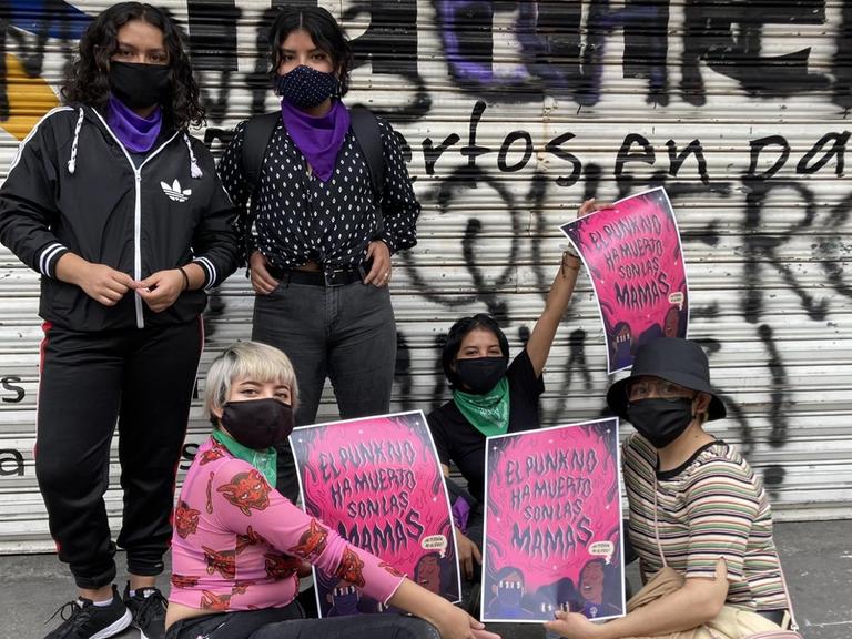 Fünf junge Frauen mit Masken und Plakaten stehen und sitzen auf der Straße vor einem Gebäude mit geschlossenen Rollos, die mit Graffiti besprüht sind.