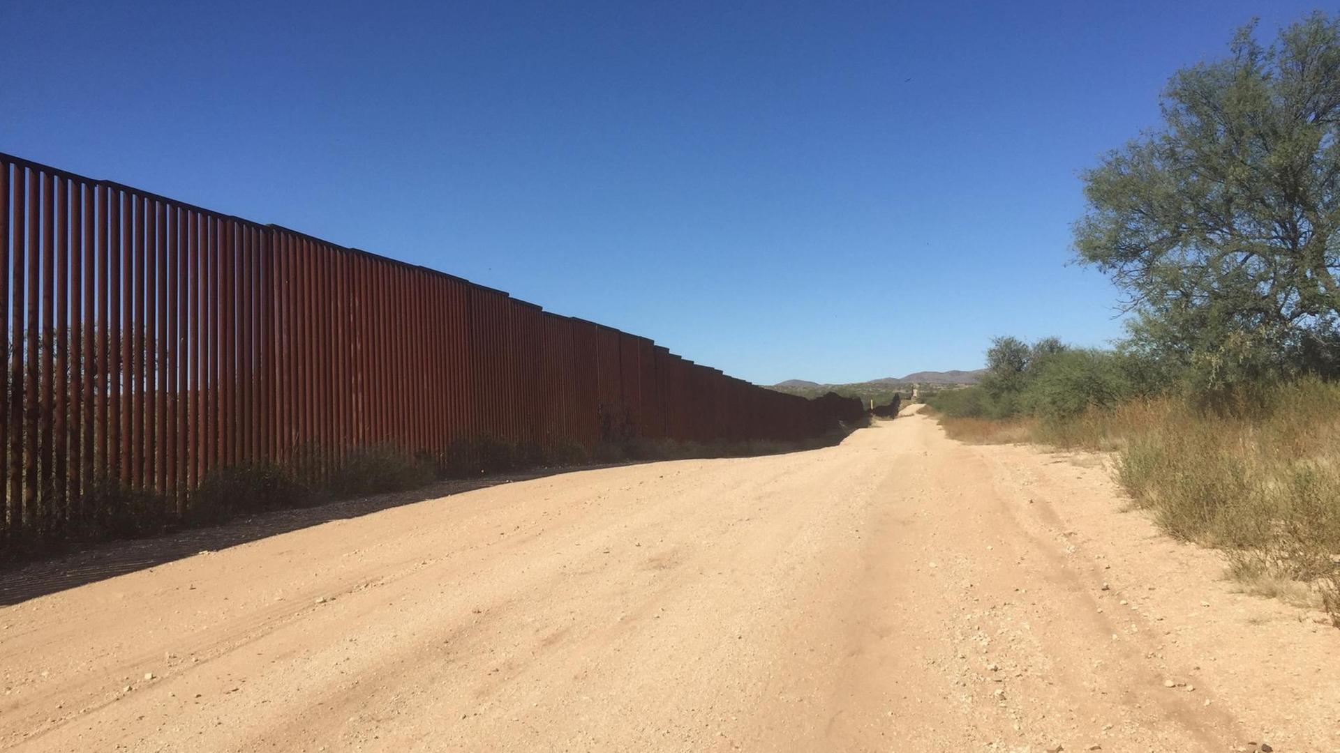 Am linken Bildrand sieht man den Grenzzaun zwischen Mexiko und den USA. In der Bildmitte verläuft ein sandiger Weg und am rechten Bildrand sind karge Bäume und Büsche zu sehen.