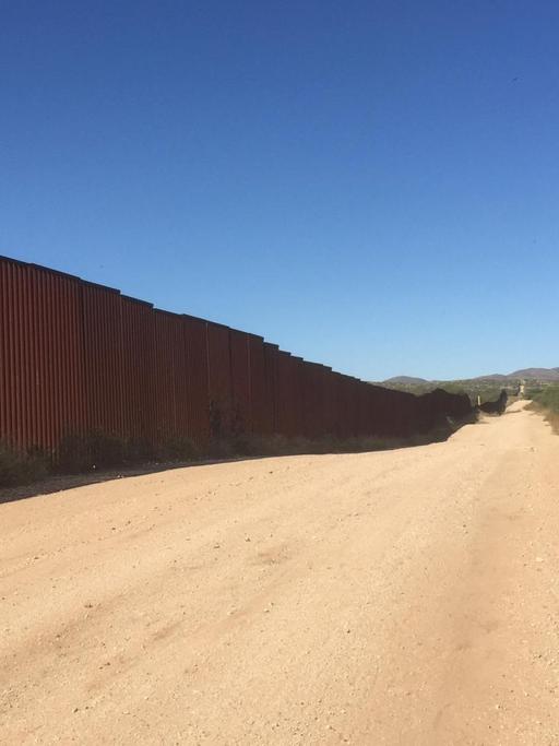 Am linken Bildrand sieht man den Grenzzaun zwischen Mexiko und den USA. In der Bildmitte verläuft ein sandiger Weg und am rechten Bildrand sind karge Bäume und Büsche zu sehen.