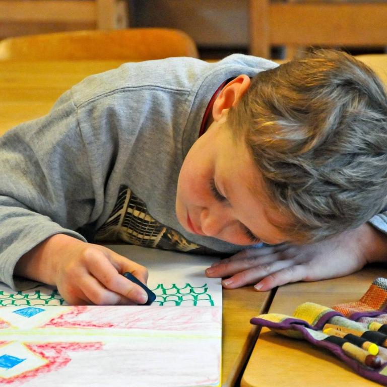 Ein circa achtjähriger blonder Junge hängt über ein Bild gebeugt und malt eifrig mit Kreiden. Er befindet sich in einer Waldorfschule.