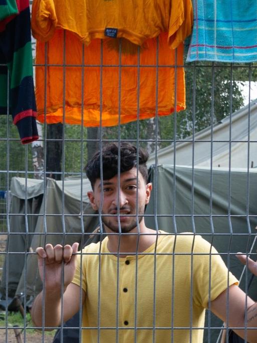 Ein junger Mann steht in einem Flüchtlingslager hinter einem Zaun.