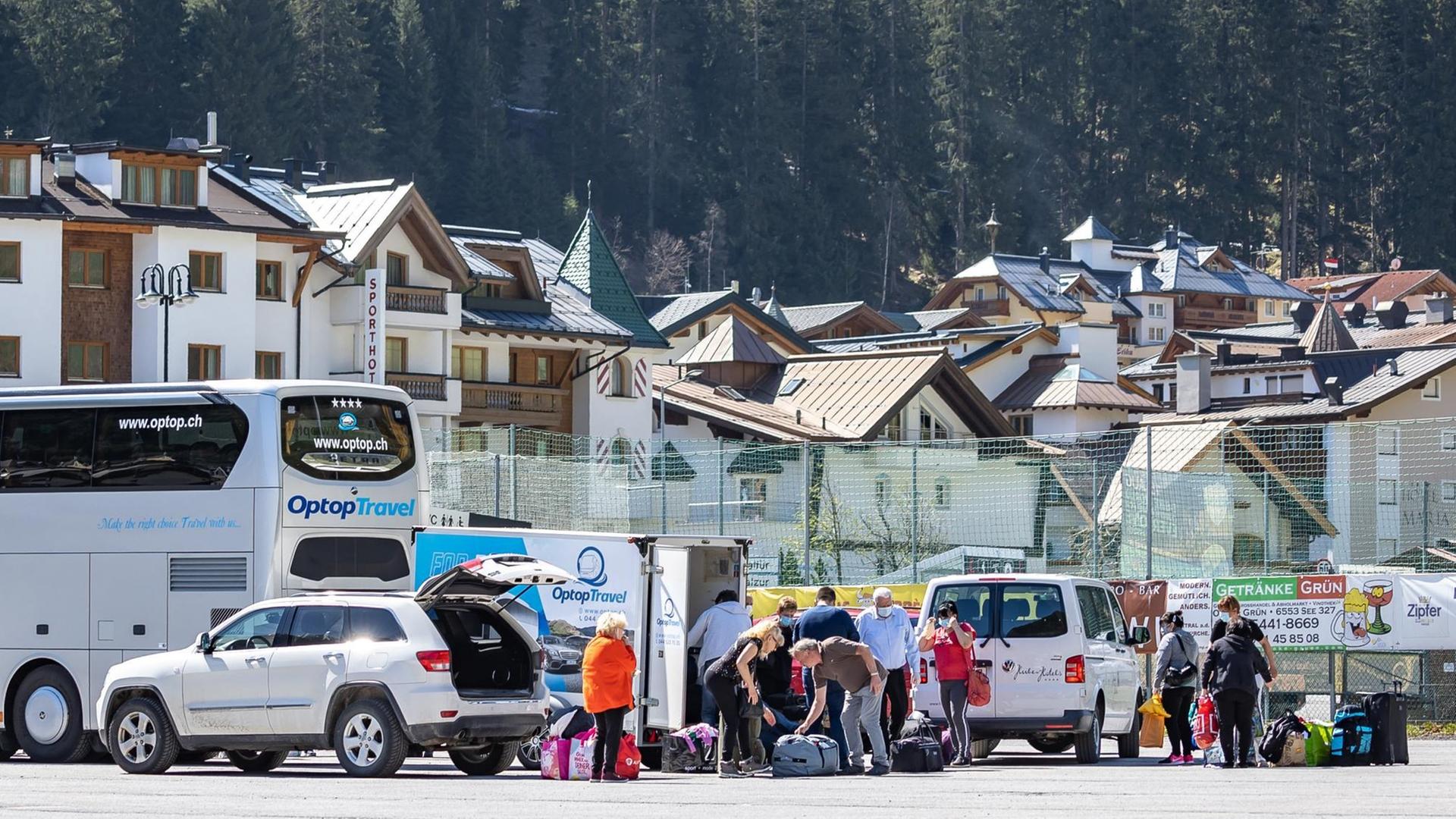 Eine Gruppe von Menschen ist mit Gepäck an einem Parkplatz um Autos und einen Bus versammelt, im Hintergrund ist die Häuswersilhouette von Ischgl zu sehen.