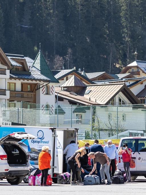 Eine Gruppe von Menschen ist mit Gepäck an einem Parkplatz um Autos und einen Bus versammelt, im Hintergrund ist die Häuswersilhouette von Ischgl zu sehen.