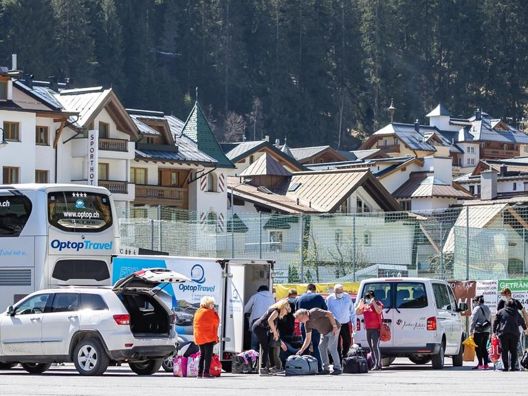 Eine Gruppe von Menschen ist mit Gepäck an einem Parkplatz um Autos und einen Bus versammelt, im Hintergrund ist die Häuswersilhouette von Ischgl zu sehen.