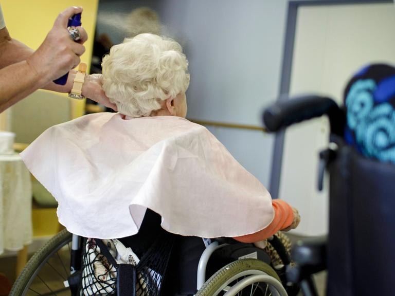 Eine Friseurin frisiert in einem eine alte Frau, die im Rollstuhl sitzt.