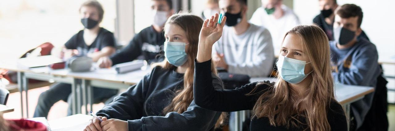Schülerinnen und Schüler nehmen mit Mund- und Nasenschutz am Unterricht teil, eine Schülerin meldet sich.