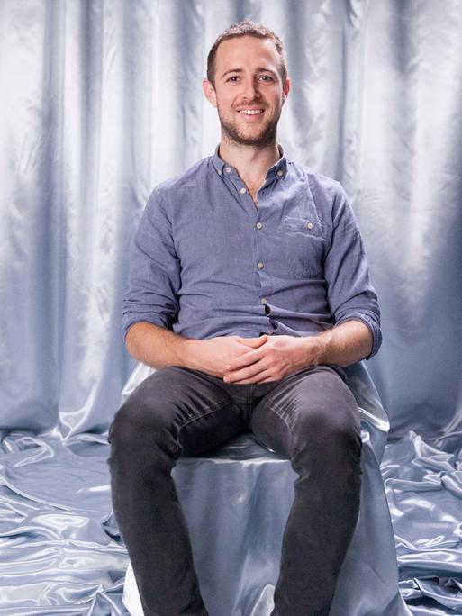 Auf einem Stuhl, der genau wie der Boden und der Hintergrund mit einem silberfarbenen Stoff abgedeckt ist, sitzt Interviewpartner Lukas in dunkler Jeans und blauen Jeanshemd und schaut lächelnd in die Kamera.