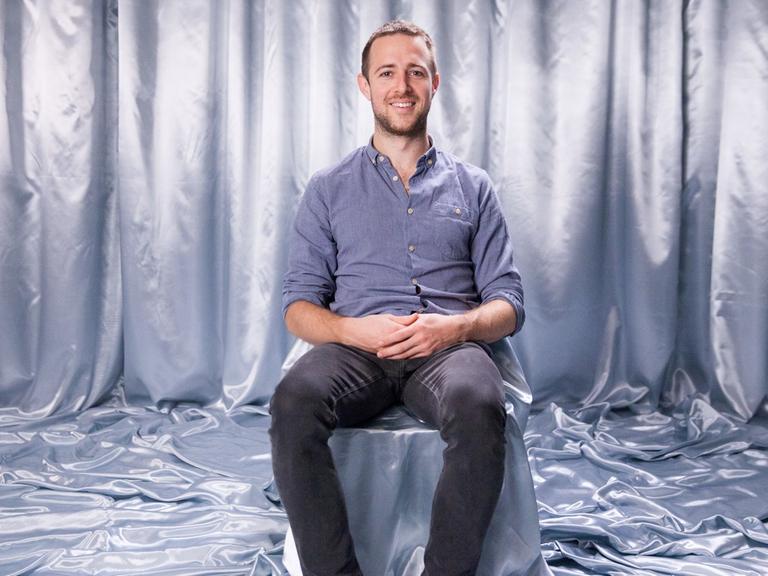 Auf einem Stuhl, der genau wie der Boden und der Hintergrund mit einem silberfarbenen Stoff abgedeckt ist, sitzt Interviewpartner Lukas in dunkler Jeans und blauen Jeanshemd und schaut lächelnd in die Kamera.