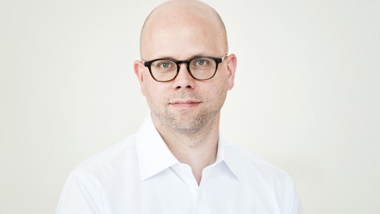 Ein Mann mit weißem Hemd und dunkler Brille schaut freundlich in die Kamera. Es ist Clemens Stachel.