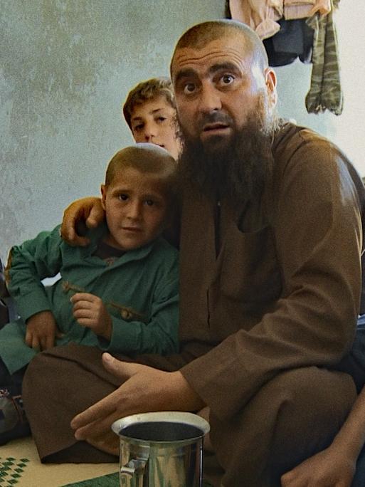 Ein Mann mit Bart und kahl geschorenem Kopf sitzt von Kindern umringt auf einer Decke in einem kargen Haus. Alle schauen aufmerksam in Richtung des Betrachters.