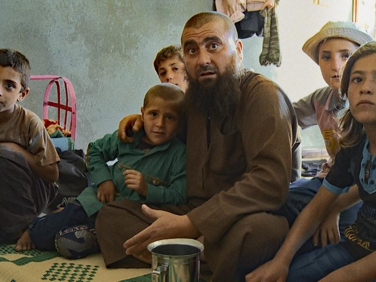Ein Mann mit Bart und kahl geschorenem Kopf sitzt von Kindern umringt auf einer Decke in einem kargen Haus. Alle schauen aufmerksam in Richtung des Betrachters.
