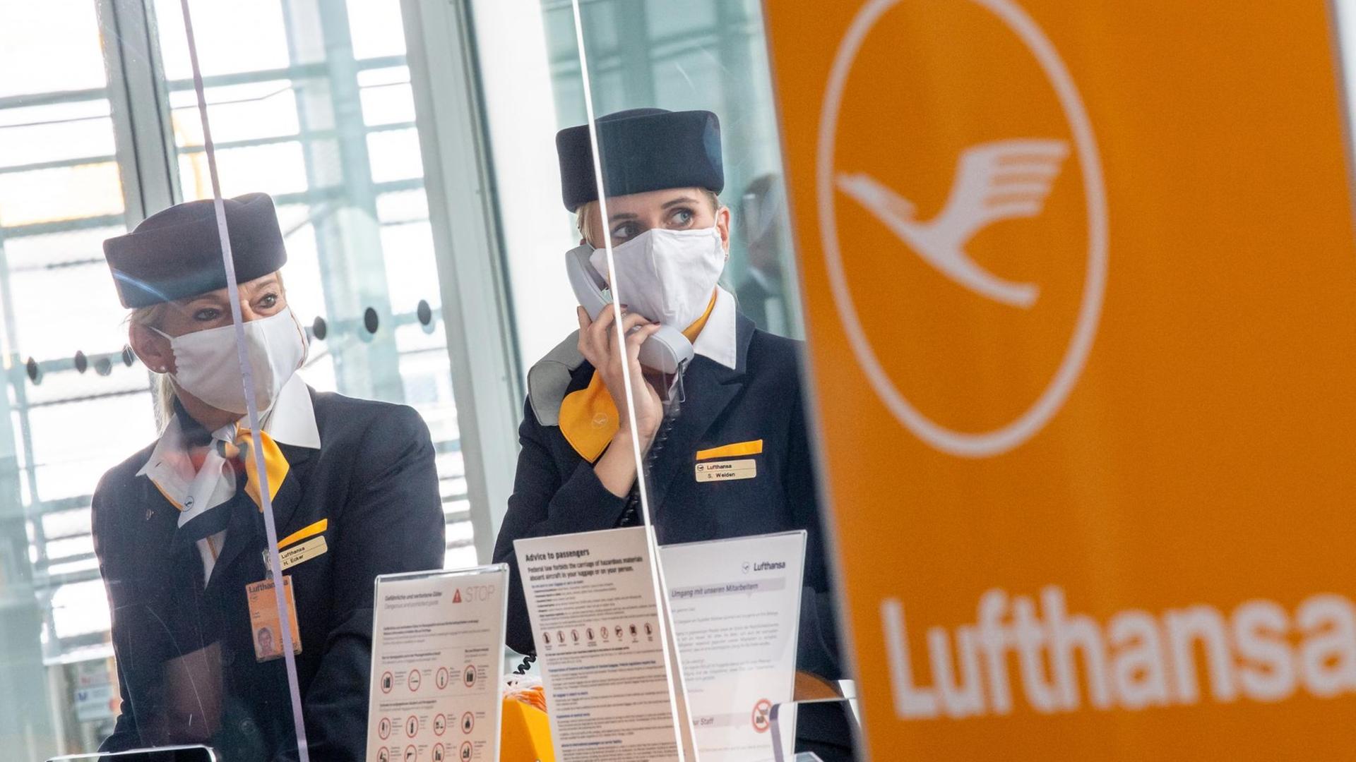 Zwei Lufthansa-Mitarbeiterinnen mit Maske und hinter Plexiglasscheiben fertigen Fluggäste ab. 