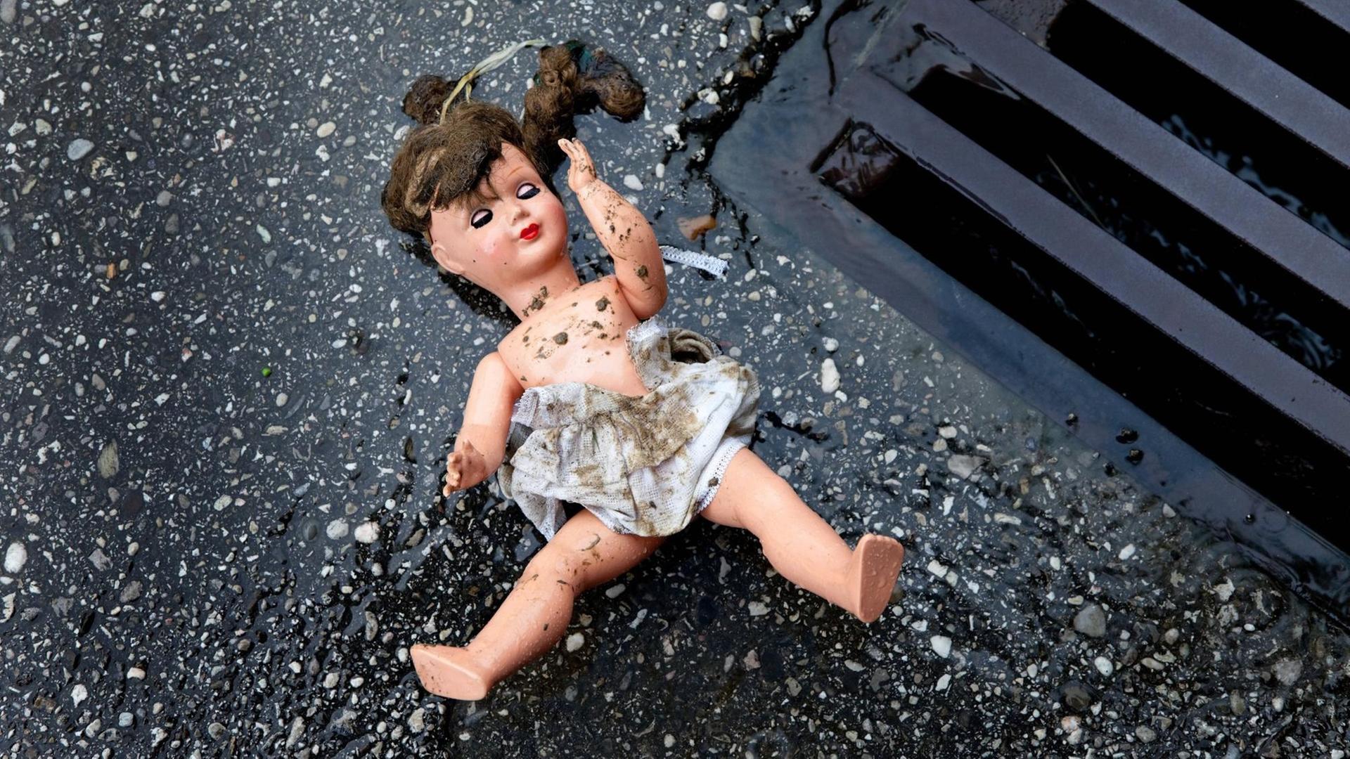 Zu sehen ist eine Puppe mit schmutzigem Kleid, die auf dem Boden neben einem Gulli liegt.