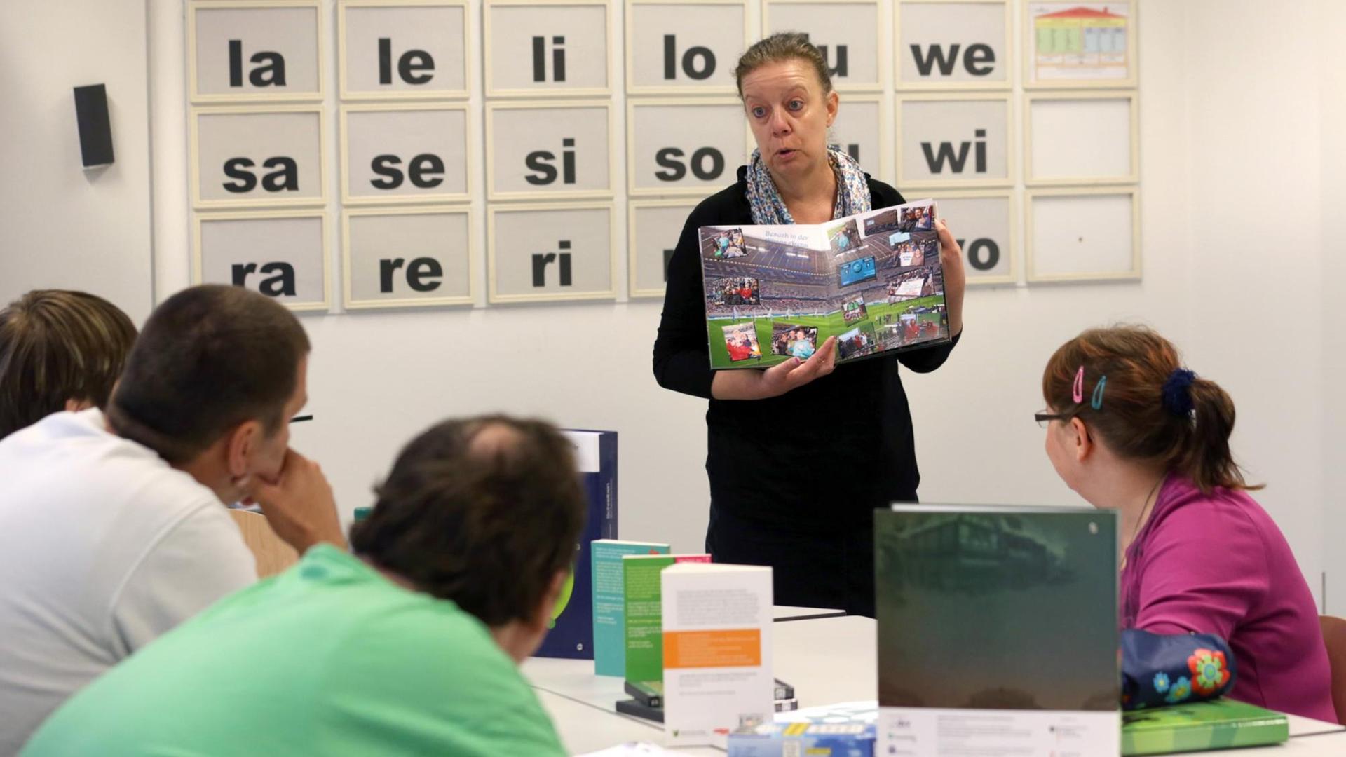 Erwachsene sitzen in einem Klassen-Zimmer und lernen lesen und schreiben. Eine Lehrerin zeigt ein Buch. An der Tafel sind Buchstaben zu lesen.