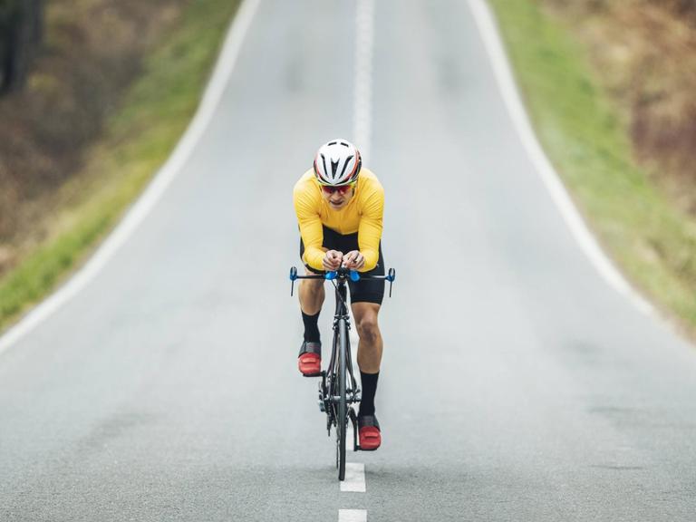 Ein Mann mit gelbem Shirt und Fahrradhelm fährt auf einer asphaltierten Strasse Rennrad
