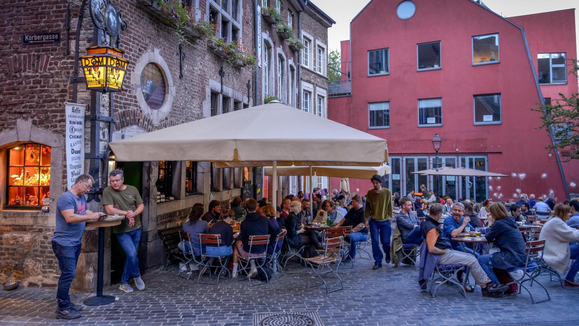 Das Bild zeigt mehrere Menschen, die draußen an Tischen eines Restaurants sitzen