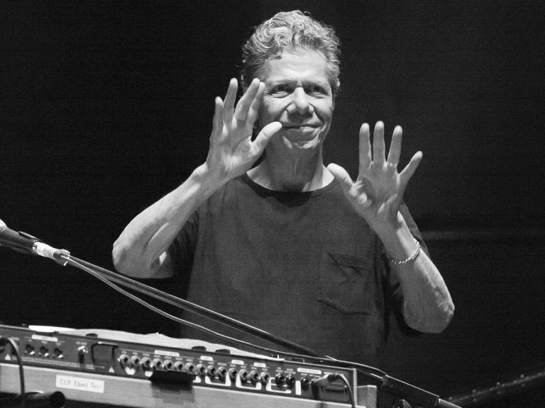 Die Schwarzweißfotografie zeigt einen Mann im T-Shirt mit grauem, weilligen Haar hinter einem E-Piano. Er hält beide Hände in die Höhe, die Handflächen zeigen wie zum Segen nach vorn.