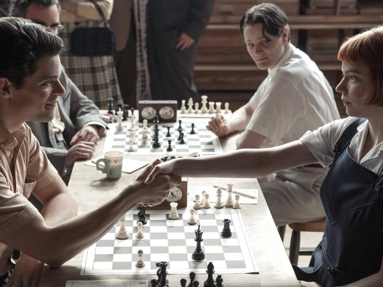 Szene aus der Netflix-Serie "Das Damengambit". Ein Mann und eine Frau sitzen vor einem Schachbrett und begrüßen sich.