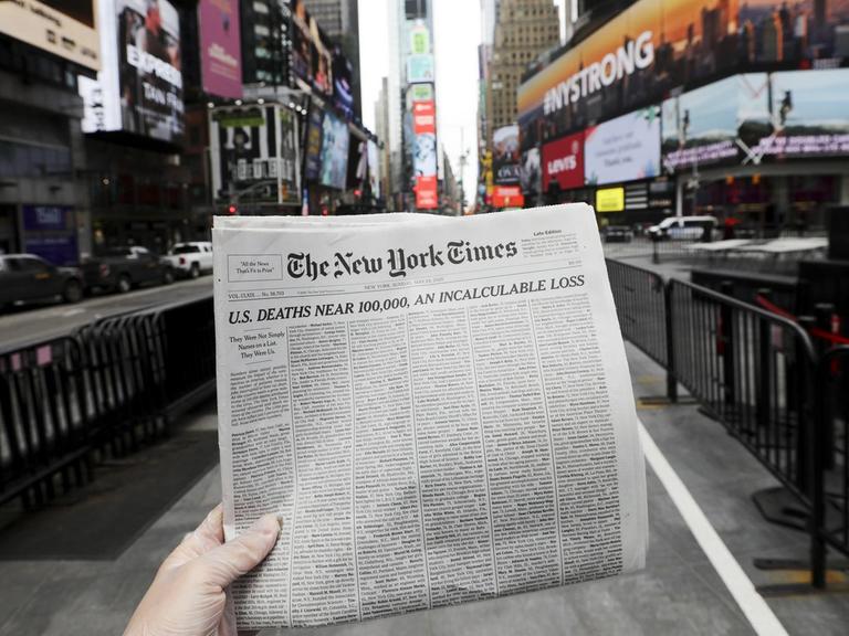 Eine Händ hält die Titelseite der New York Times, vor die Kulisse der Time Square in New York.