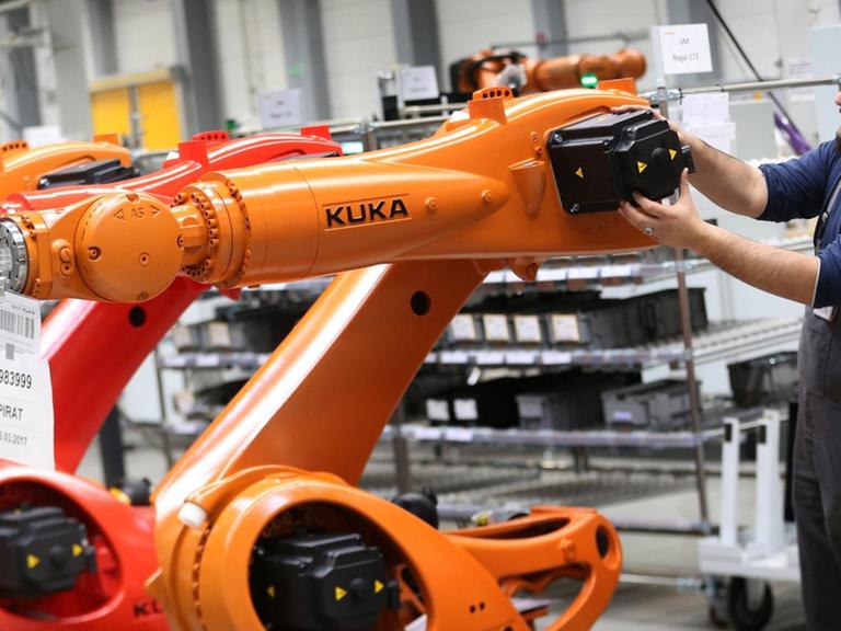 Ein Arbeiter hantiert in einer Halle an einem orangefarbenen Kuka-Roboter, daneben stehen weitere Roboter der gleichen Bauart.