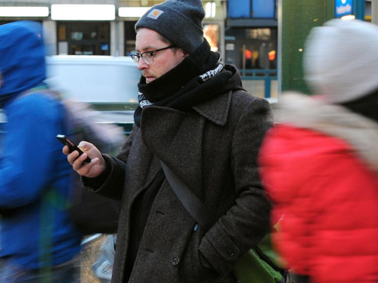 Ein junger Mann steht am 24.01.2015, mit dem Blick auf sein Smartphone gerichtet, an einer Straße in Berlin.