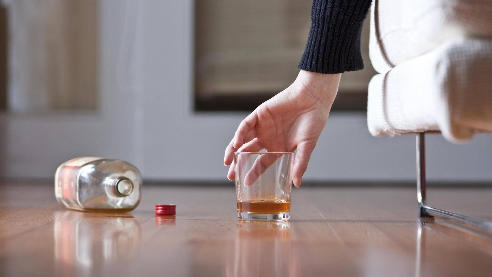 Eine Hand greift nach einer leeren Schnapsflasche, daneben steht ein Glas mit Alkohol.
