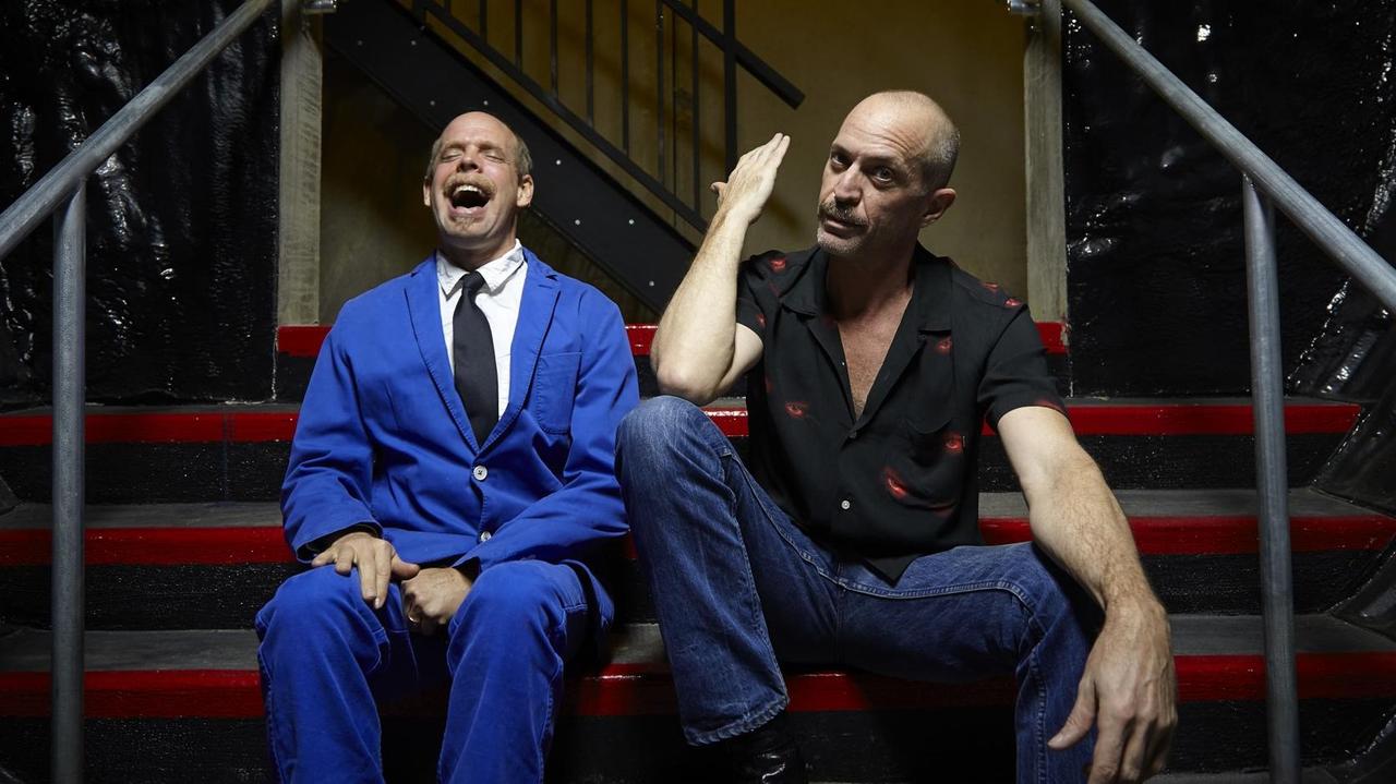 Zwei Männer sitzen auf einer Treppe. Der linke Mann im blauen Anzug lacht. Der Mann rechts stützt sein Ellenbogen auf sein Knie.