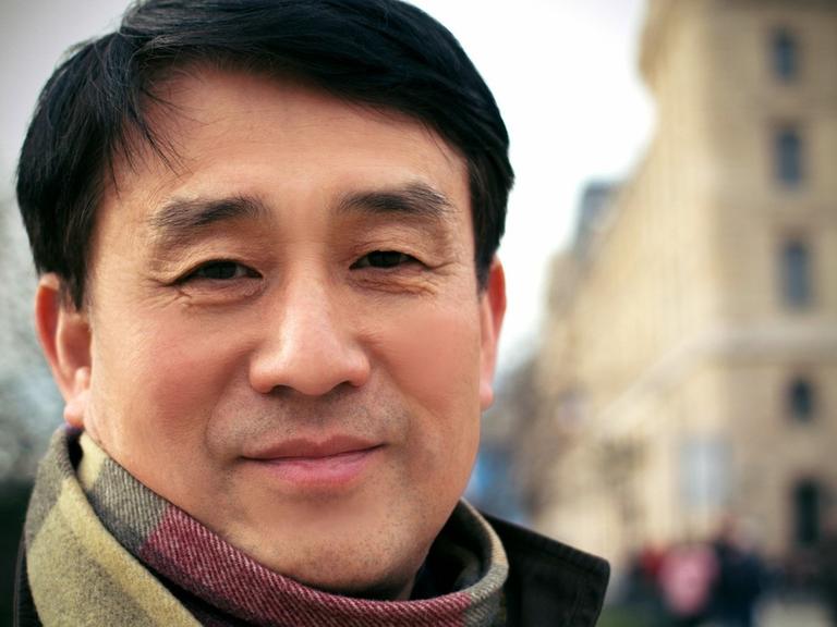 Auf dem Porträtbild ist ein lächelnder Mann mit asiatischem Aussehen vor einer Häuserzeile zu sehen.