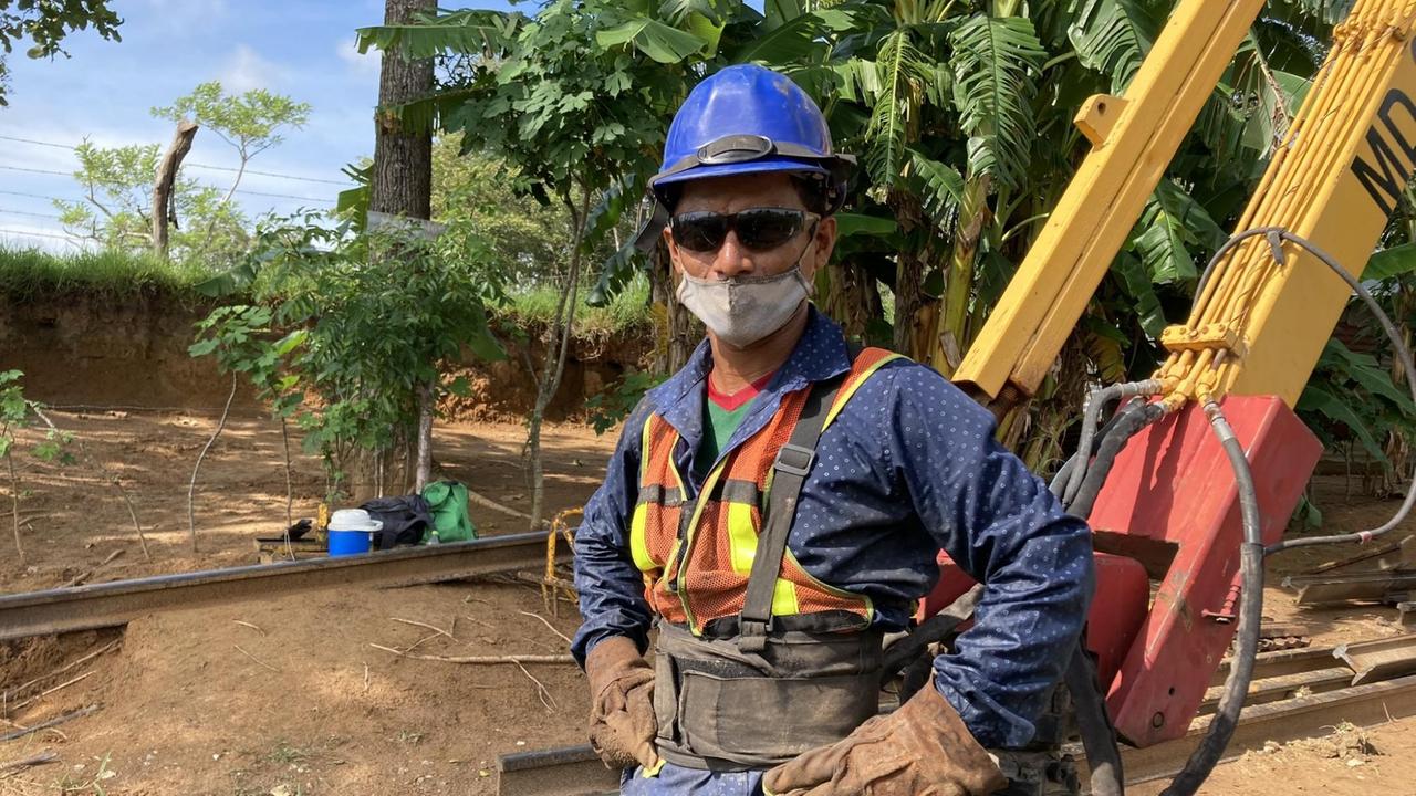 Ein Mann steht in Arbeitskleidung mit Sonnenbrille, Bauhelm und Mundschutz auf einer Baustelle.