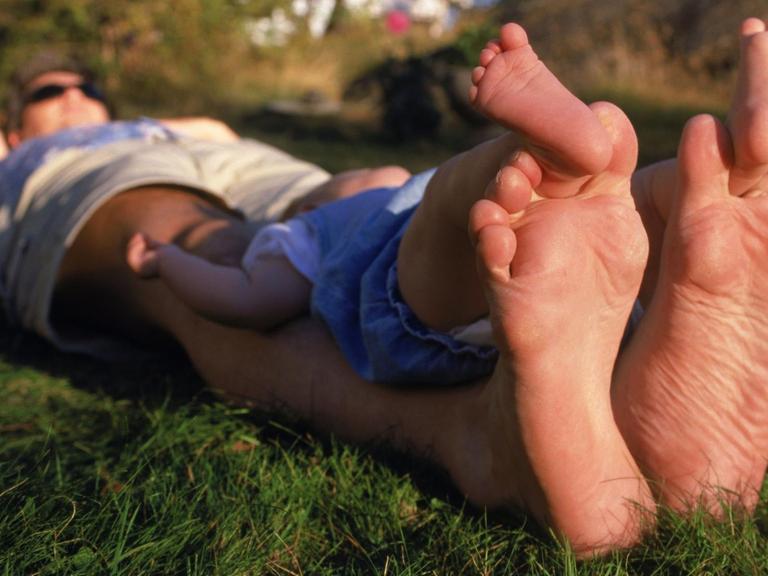 Ein Vater und Kind liegen im Gras und ruhen sich, die Füße weit von sich gestreckt, aus.