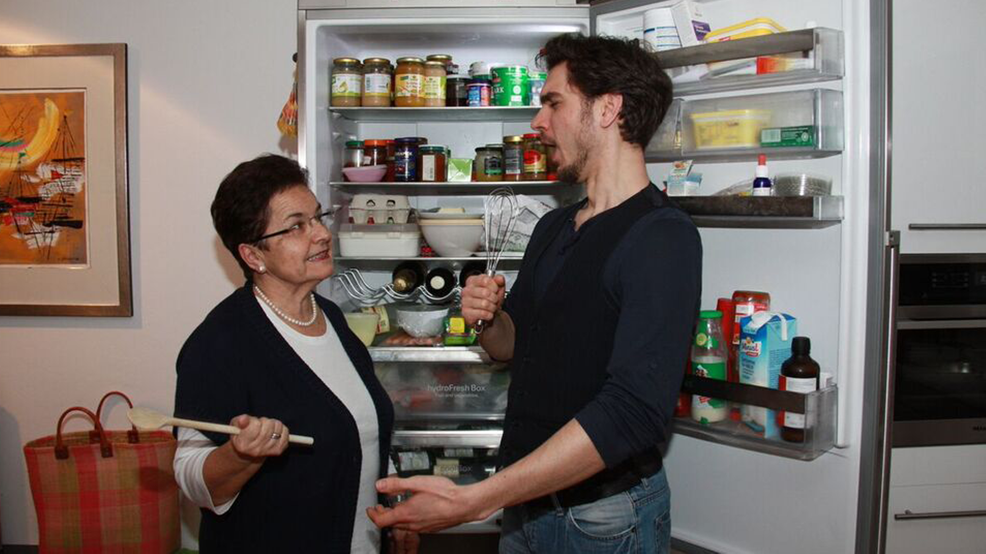 Sebastian alias Stefan Görgner steht mit seiner Mutter vor dem Kühlschrank. Beide halten Küchenutensilien in der Hand. Er singt.