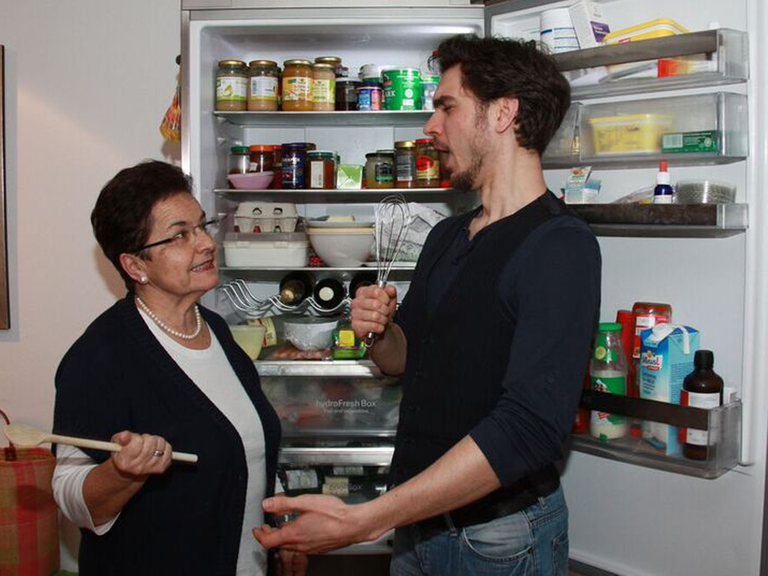 Sebastian alias Stefan Görgner steht mit seiner Mutter vor dem Kühlschrank. Beide halten Küchenutensilien in der Hand. Er singt.