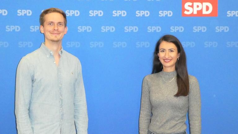 Peter Maaß und Sinem Tasan-Funke stehen vor einer Plakatwand und lächeln. Peter Maaß und Sinem Tasan-Funke stehen vor einer Plakatwand und lächeln.