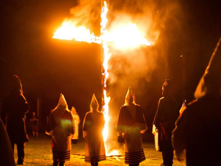 Mitglieder des Geheimbundes Ku-Klux-Klan in den USA stehen in weißen Gewändern und Kapuzen um ein brennendes Kreuz.