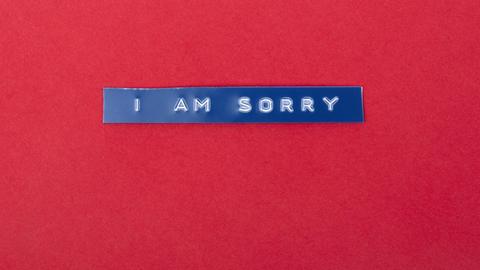 Auf rotem Untergrund liegt ein blauer Plastikstreifen auf den die Worte „I am sorry“ geprägt sind