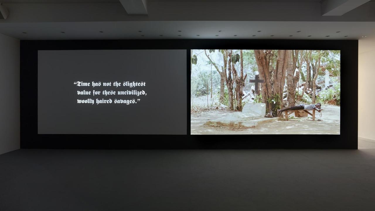 Ausstellungsansicht: Eine zweigeteilte Videoprojektion: Rechts ein Junge neben einem Grab, links ein Satz in Frakturschrift.