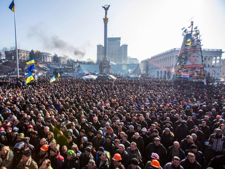 Eine große Menschenmenge auf dem Maidan-Platz in Kiew, im Hintergrund eine ukrainische Flagge an einem Mast, das Monument in der Mitte des Platzes und das von den Demonstranten errichtete Mahnmal mit dem Bild der inhaftierten Ex-Regierungschefin Julia Timoschenko