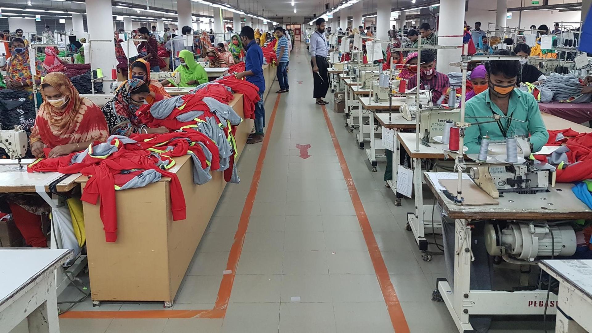 Ein Blick in eine Textilfabrik in Bangladesch. Ein Gang befindet sich zwischen einer Reihe von Tischen mit Nähmaschinen an denen Frauen sitzen und Kleidung herstellen.